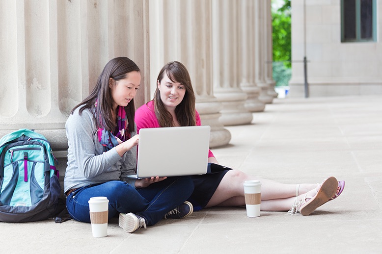 two female students studying outdoors on university campus with laptop computer